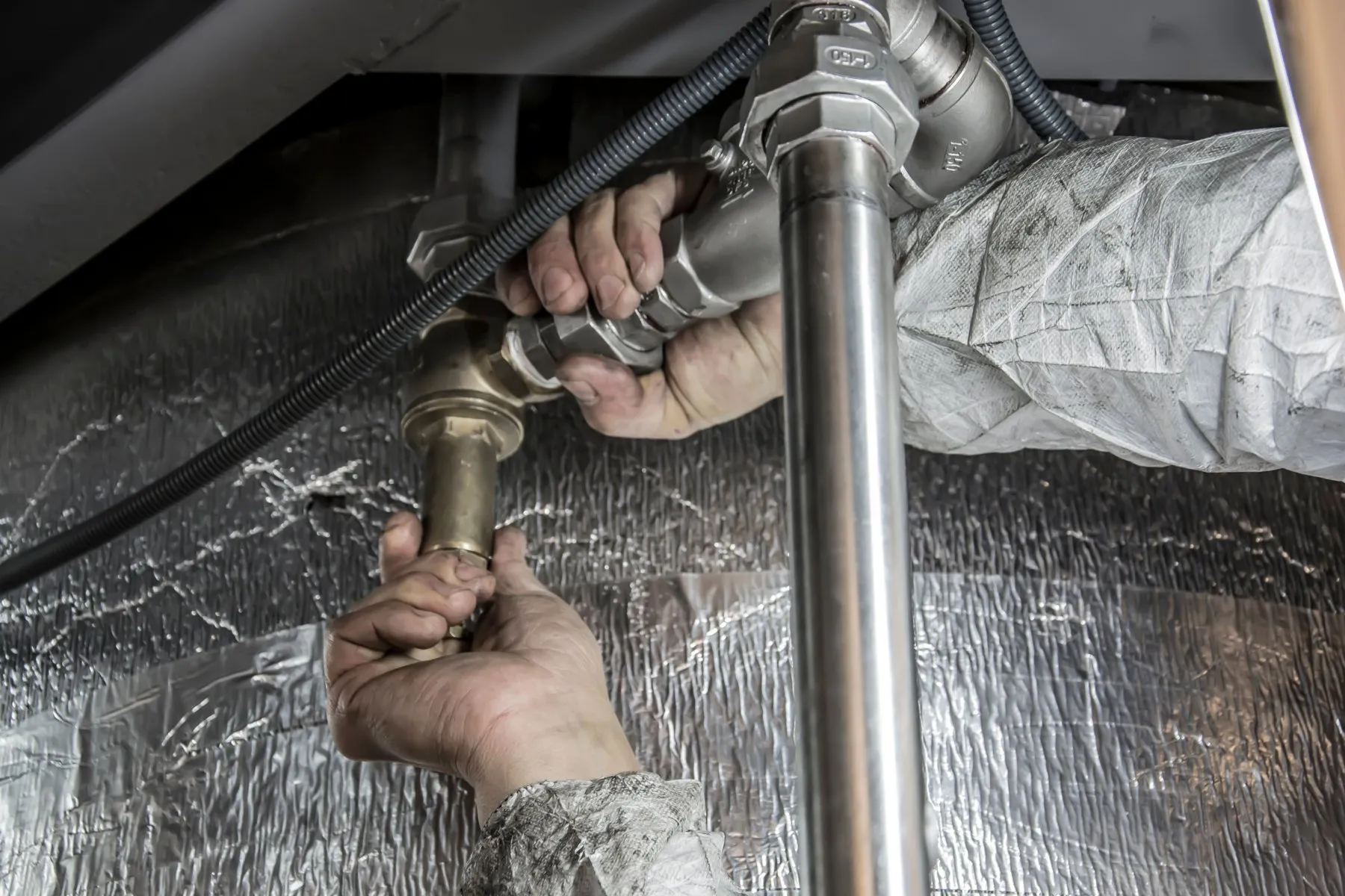A plumber's hands tightening a brass valve on steel pipework under a floor